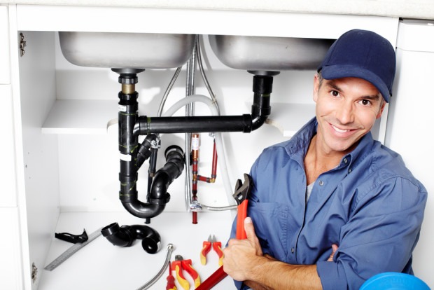 A plumber in a blue uniform and cap kneels by an open kitchen sink cabinet with plumbing tools and pipes visible.