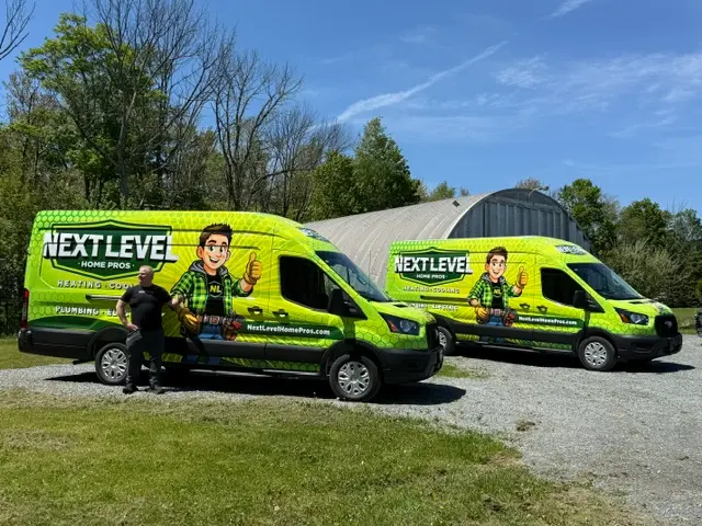 Two green Next Level Home Pros vans with cartoon graphics are parked on gravel near a metal building; a person stands beside one van.